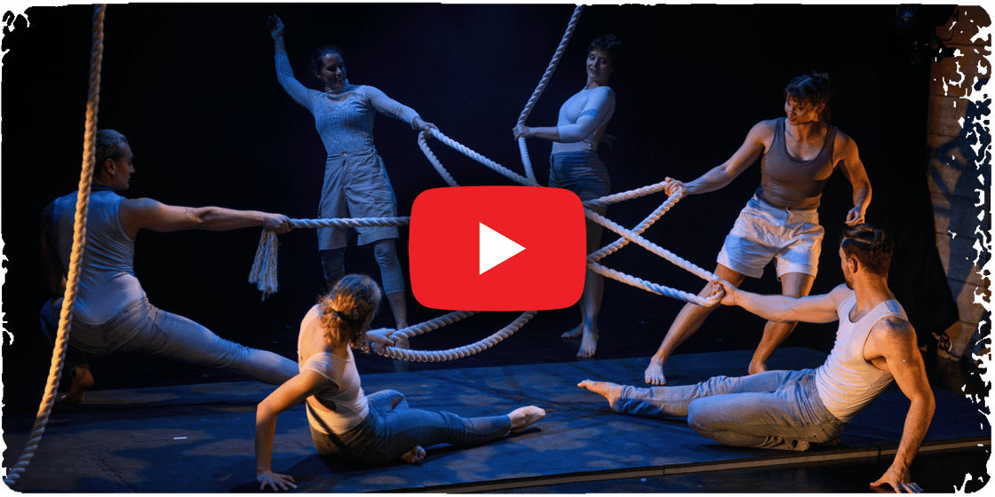 A cast of Fringe performers pull on ropes onstage. A YouTube-style 'play' button is superimposed over the centre of the image.